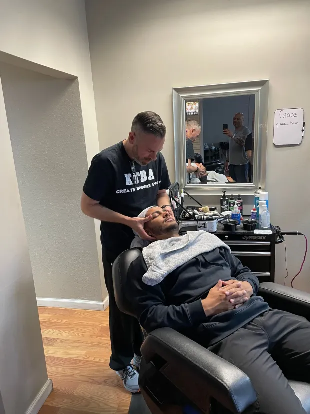 A barber adjusts a client's head position while they sit in a salon chair with a towel draped over their shoulders.