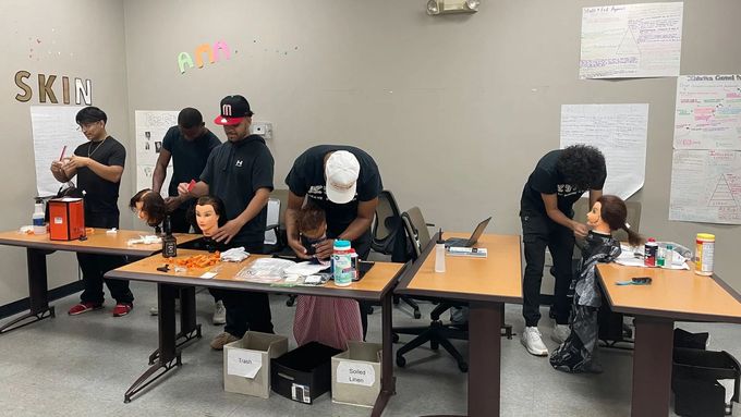 Students practice haircutting techniques on mannequin heads at desks in a classroom.