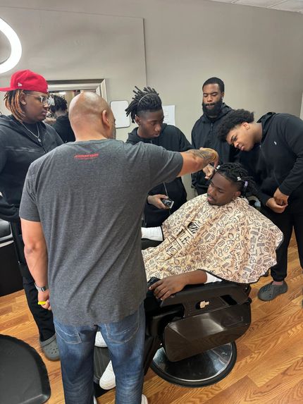 A barber cutting a client's hair in a salon while a group of people watch closely.