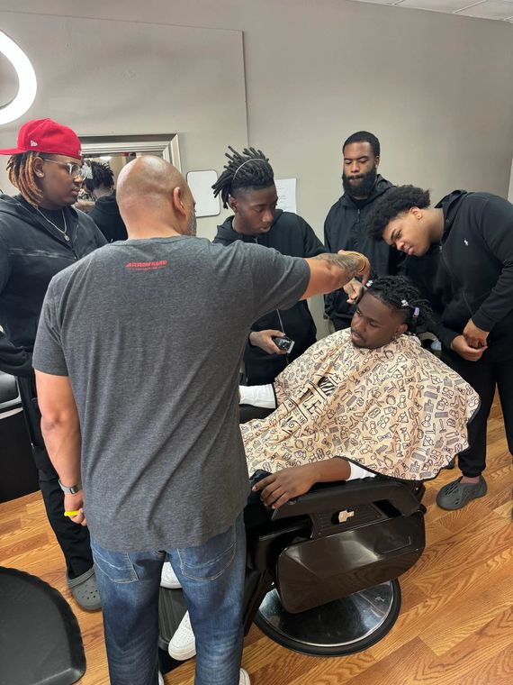 A barber cutting a client's hair in a salon while a group of people watch closely.