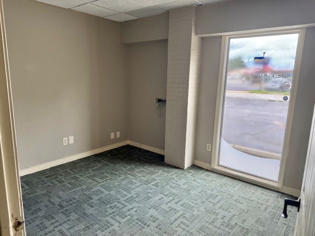 Empty office space with green carpet, gray walls, and a large window overlooking an exterior view.