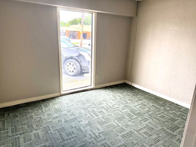 An empty room with green patterned carpet, gray walls, and a window looking out onto a street with a parked car.