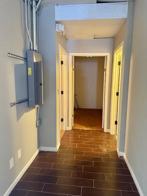 A narrow hallway with dark wood-look tile flooring and three doorways. Electrical panel and conduits on the left wall.