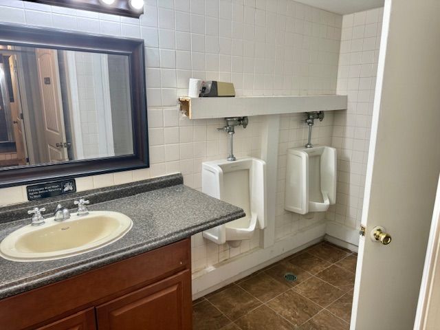 Public restroom with two urinals, a sink, and a mirror. The walls are tiled, and the floor is a brown tile.