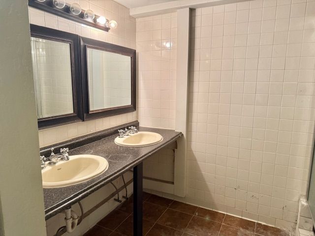 A bathroom with two sinks, mirrors, and a tiled shower, featuring dark counters and walls. The floor is brown tiles.