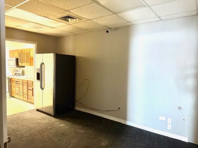 An empty office space with a refrigerator. A kitchen is visible through an open doorway.