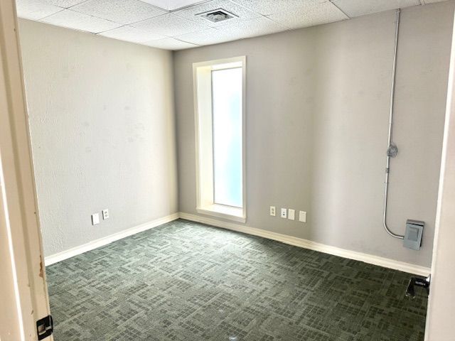 Empty office space with gray walls, green carpet, and a tall, narrow window. Electrical outlets and a cable are visible.