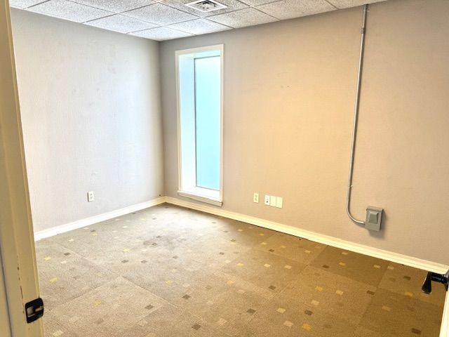 Empty room with tan walls, a tall narrow window, and patterned carpet. Electrical conduit runs up the wall.