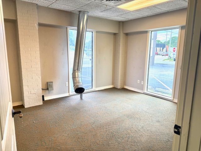 An empty, beige-walled room with a bent metal ventilation pipe hanging from the ceiling. Two large windows and a closed door are visible.