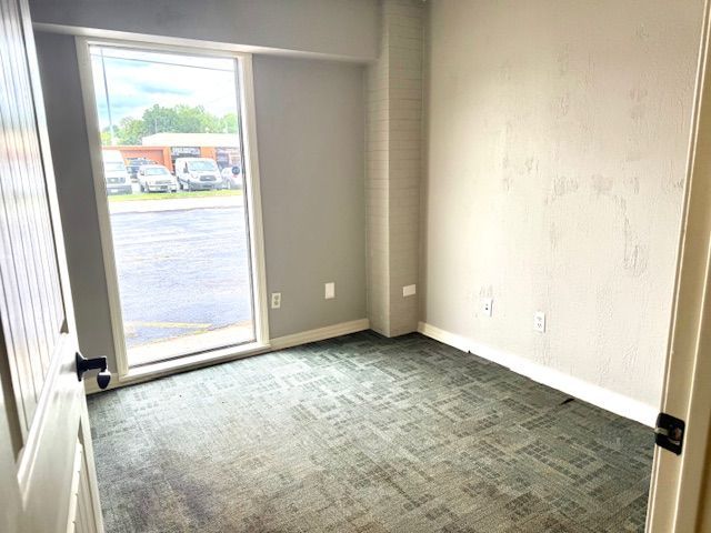 Empty office room with a large window overlooking a parking lot. Gray walls, green carpet, and a partially open white door.