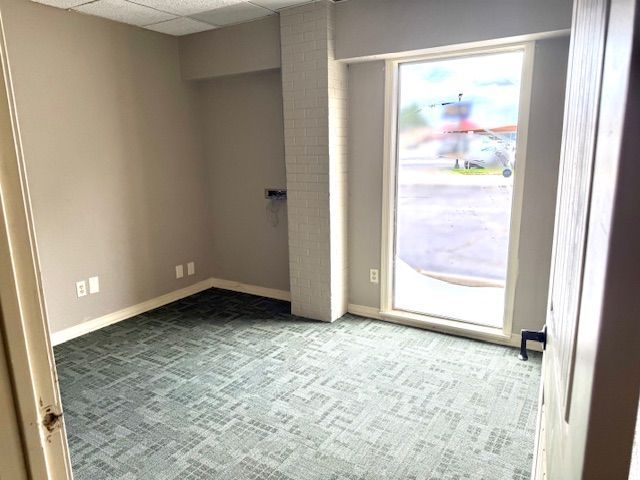 Empty office space with gray walls, green carpet, a large window, and open doorway.