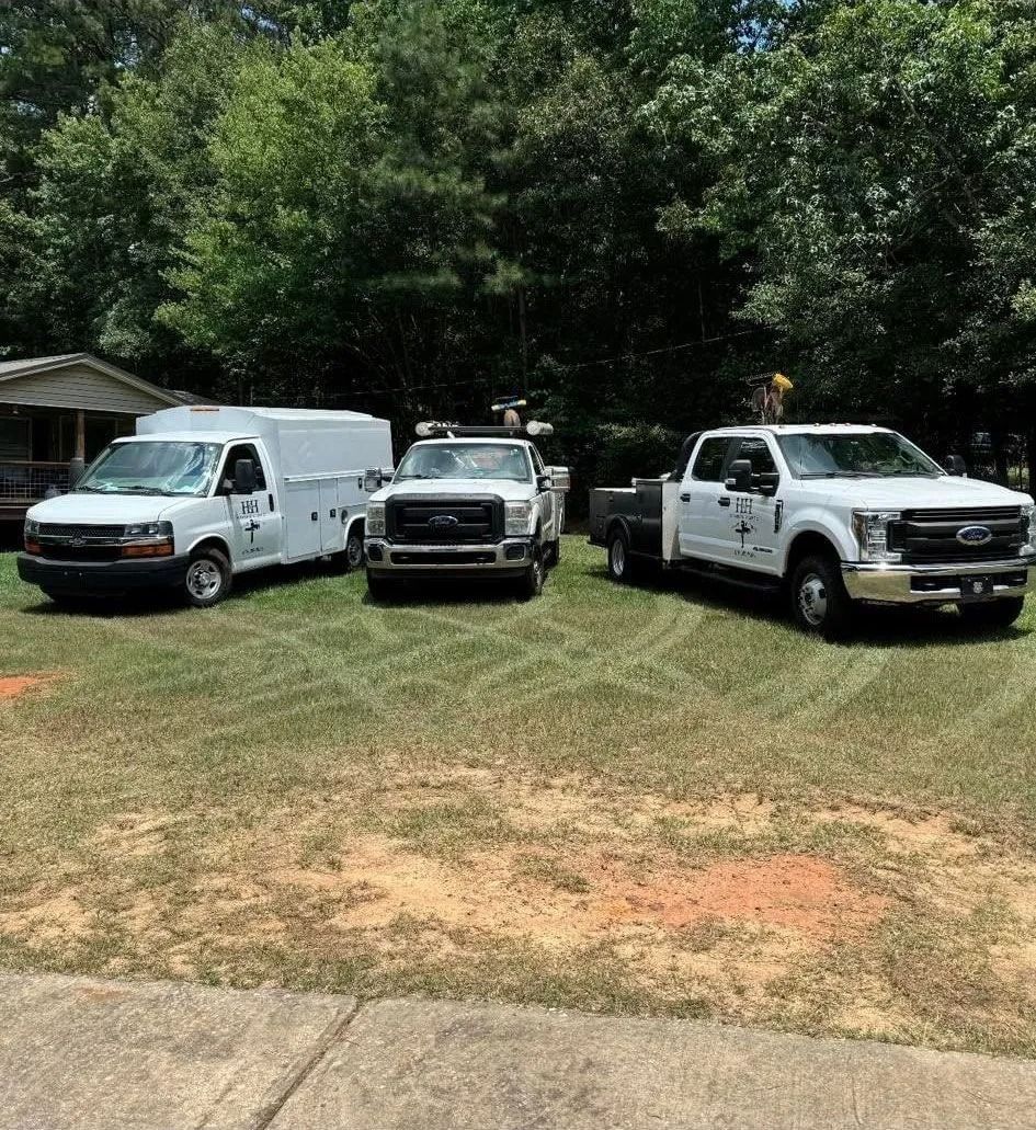 Three white work trucks parked on grass in front of trees and a building.