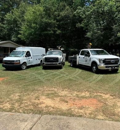 Three white work trucks parked on grass in front of trees and a building.