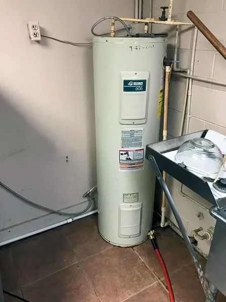 A tall, white water heater in a corner next to a wall. Red hose runs from the bottom.