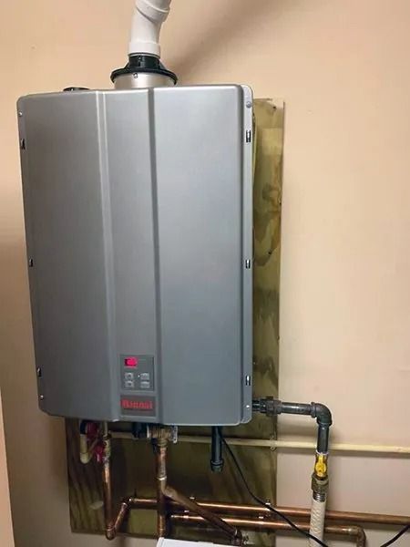 A wall-mounted, silver tankless water heater with copper pipes connected. The unit has a digital display and a white vent pipe.