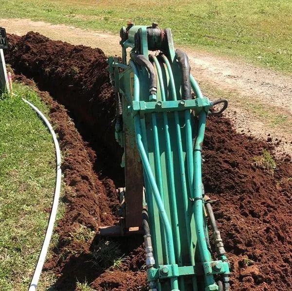 Green excavator digging a trench beside a white pipe on grass and dirt.