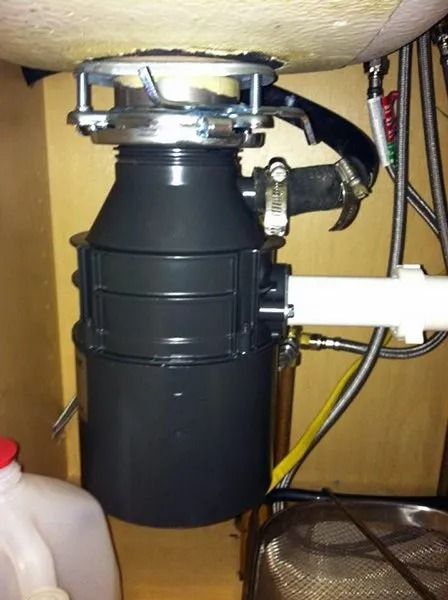 Garbage disposal unit under a kitchen sink. Black, cylindrical device with metal components and attached plumbing.