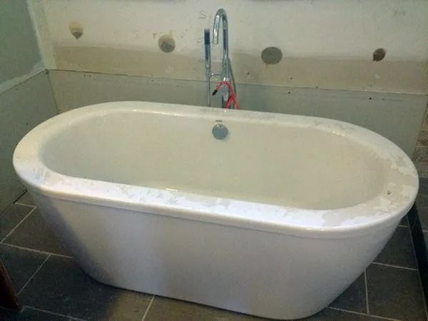 White, oval bathtub with faucet against a wall with holes, installed on tiled floor.