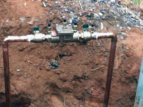 Water meter assembly installed in a shallow dirt trench with copper pipes and various valves.