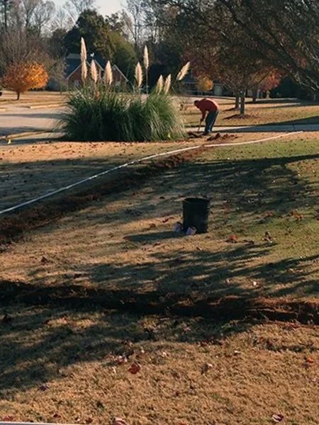 Man working near a grass area. Pampas grass in the background. Brown grass and a trash can are in the foreground.