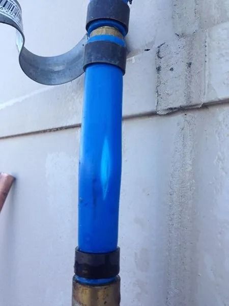 Blue PEX water pipe with a bulge, connected by brass fittings and secured by a metal strap to a wall.