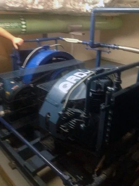 Two blue industrial generators on a metal frame, inside a room. A person's hand reaches toward one.