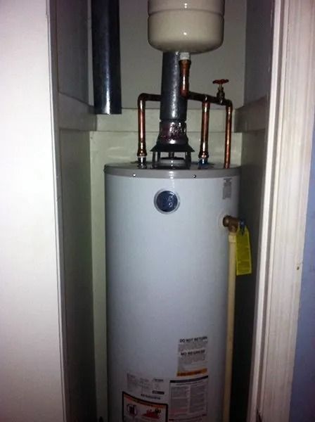 White water heater in a narrow closet with pipes and a small expansion tank above it