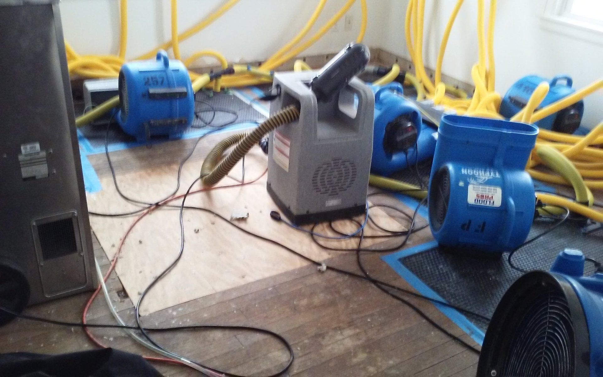 Room with drying equipment: blue air movers, hoses, and a central dehumidifier on a wet floor, indicating water damage.