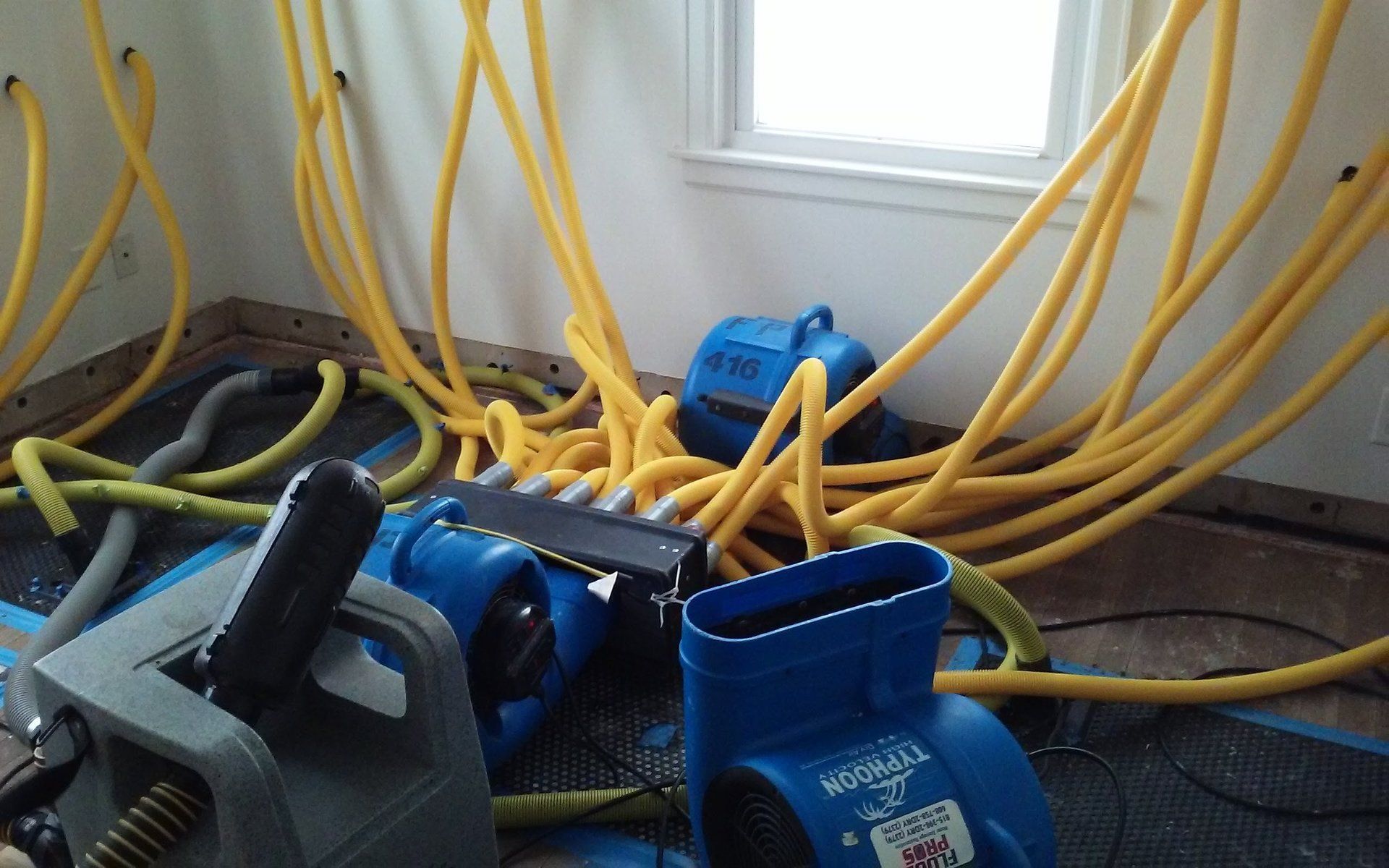 A room with water damage remediation equipment: blue air movers, yellow hoses, and a window, indicating a drying process.