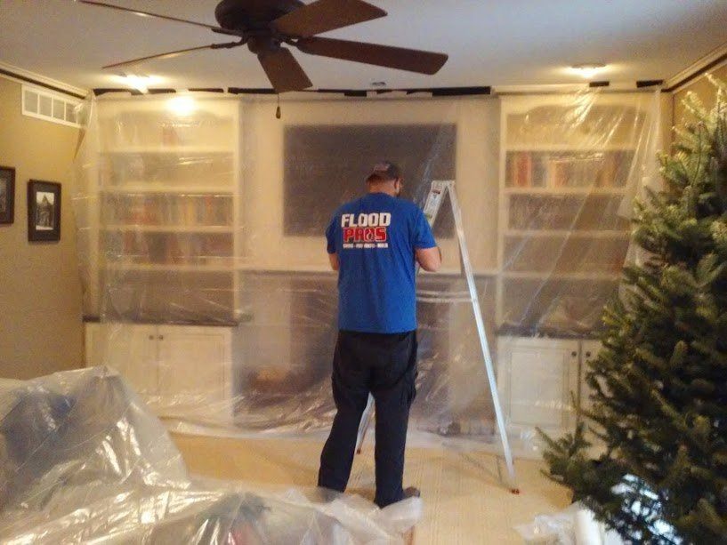 A worker in a blue shirt, standing on a ladder, works on a home entertainment system, covered in plastic sheeting.