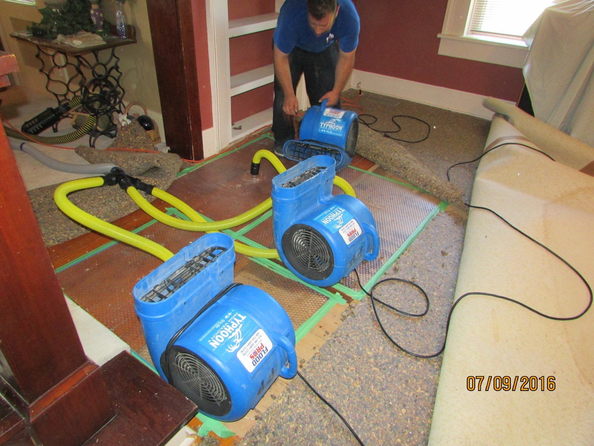 Man operates three blue air movers on a damp floor, likely for water damage restoration. Setting is an interior room with carpet and built-in shelves.