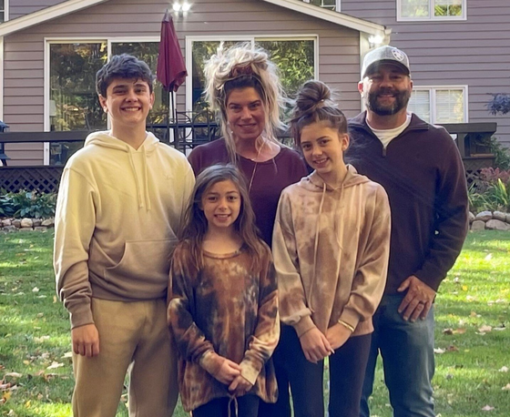 Family of five poses in front of a house; includes two teen boys, two girls, and two adults, all smiling.