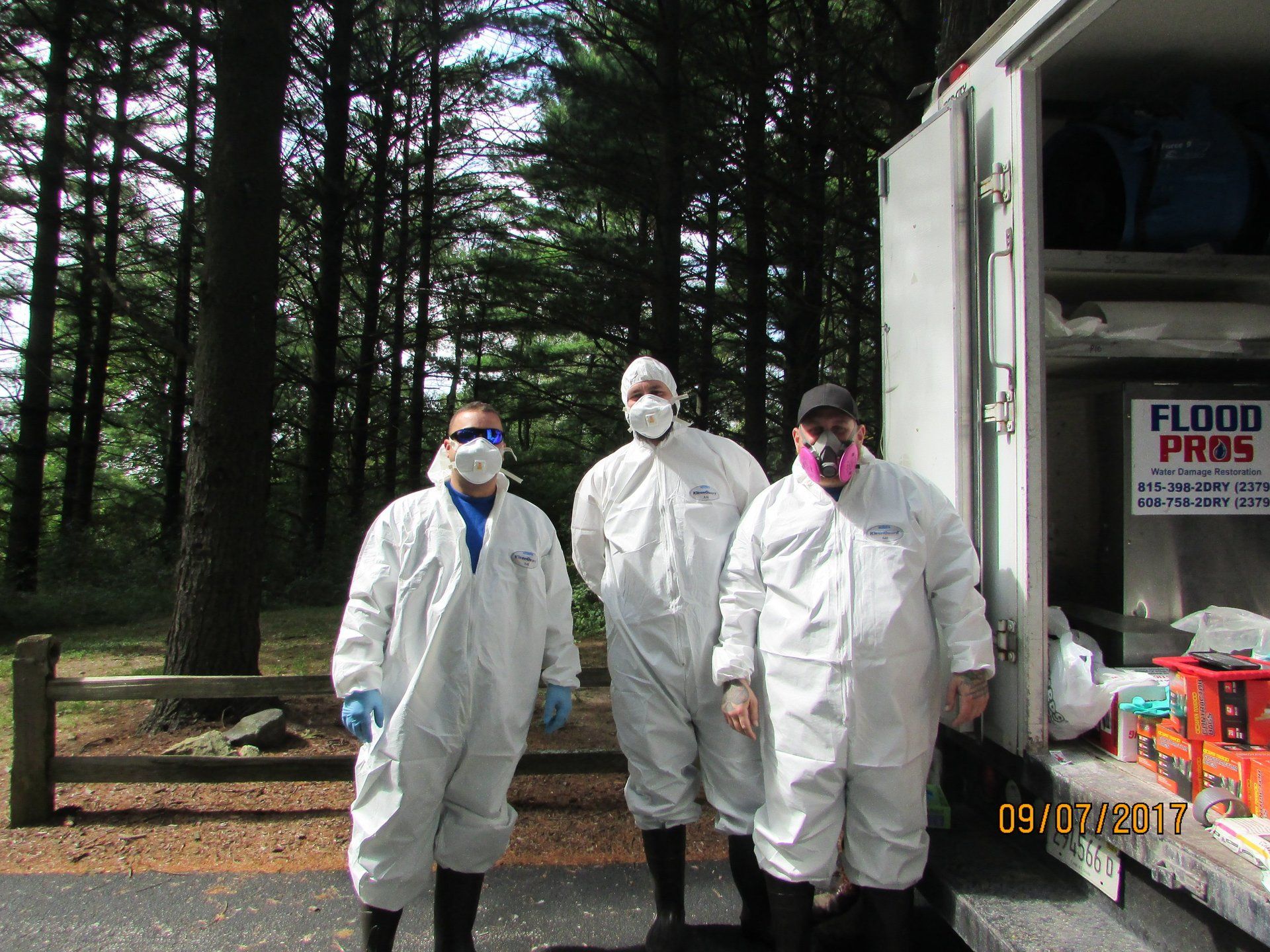 Three people in white hazmat suits stand near a truck in a wooded area; one wears a pink face mask.