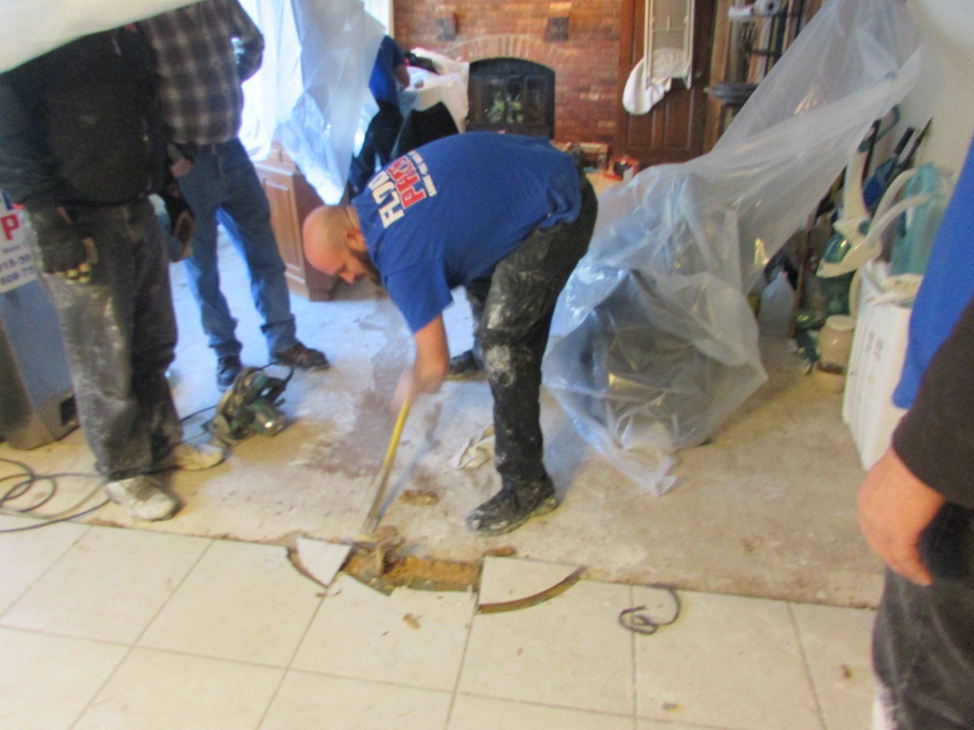 A bald man in a blue shirt scrapes at a tile floor with a tool; others watch. Construction setting.
