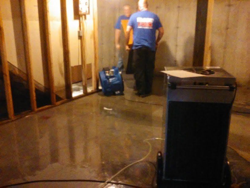 Two men in a basement cleaning up water, one operating a blue water extraction machine, the other standing nearby. The room has wet concrete floors.