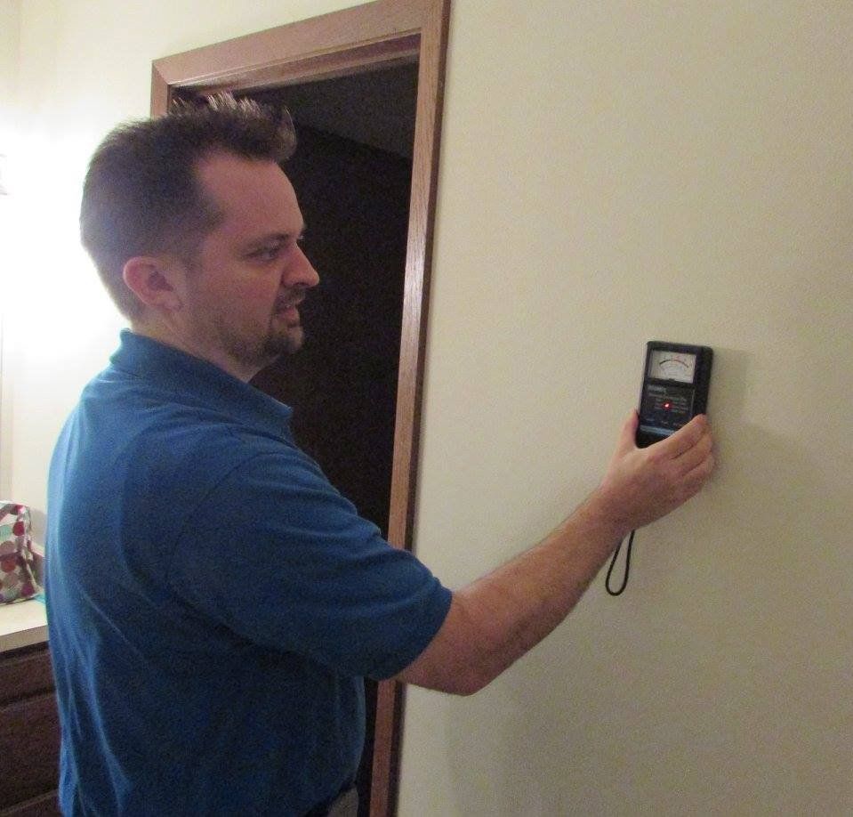 A man in a blue shirt uses a moisture meter on a white wall near a doorway.