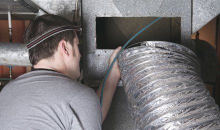 Man in grey shirt inspecting HVAC ductwork with a camera, using a headlamp for visibility.