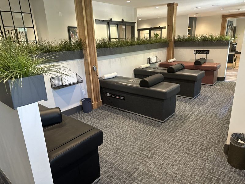 Room with several black massage tables. Gray carpet, white walls, and decorative plants.