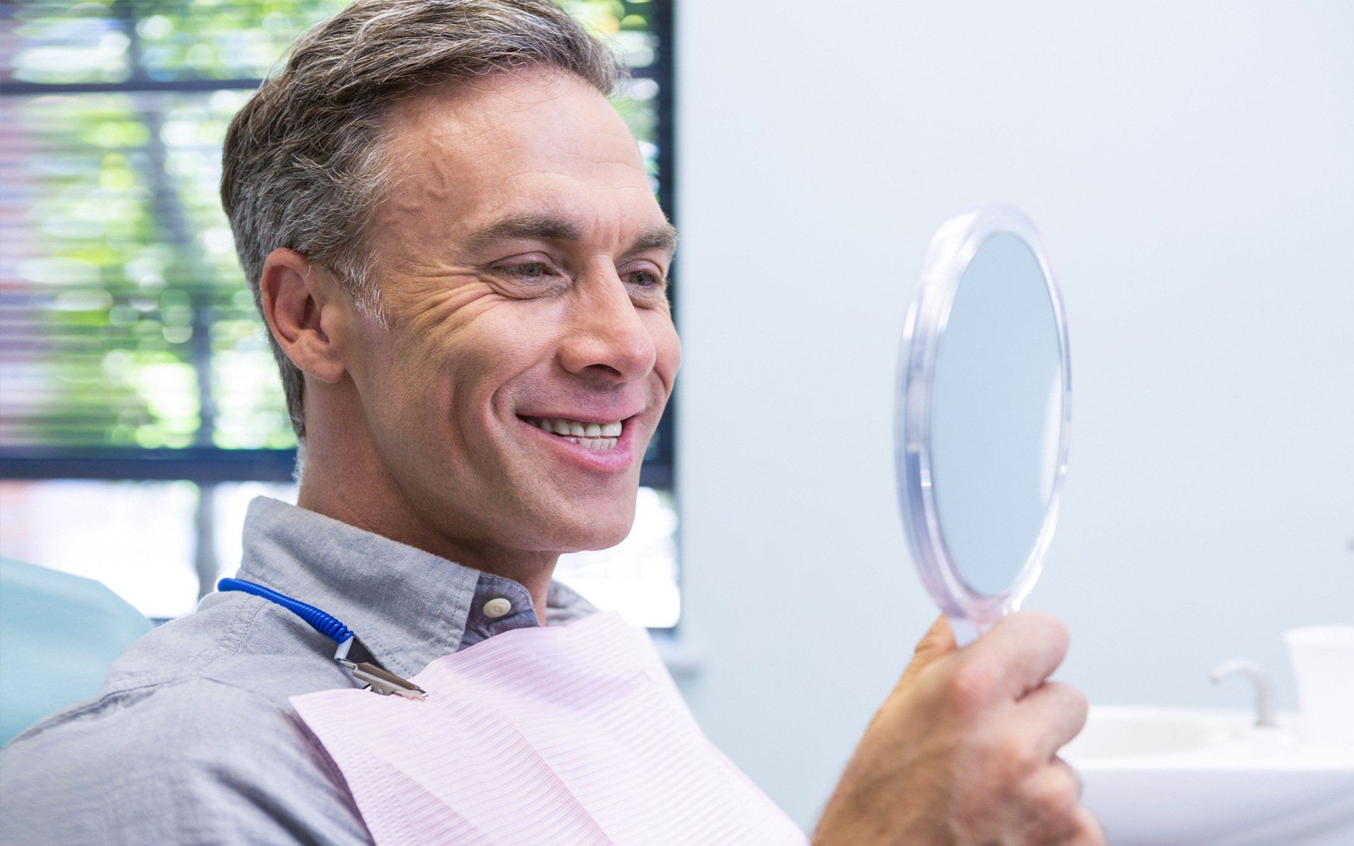 A man is sitting in a dental chair looking at his teeth in a mirror.