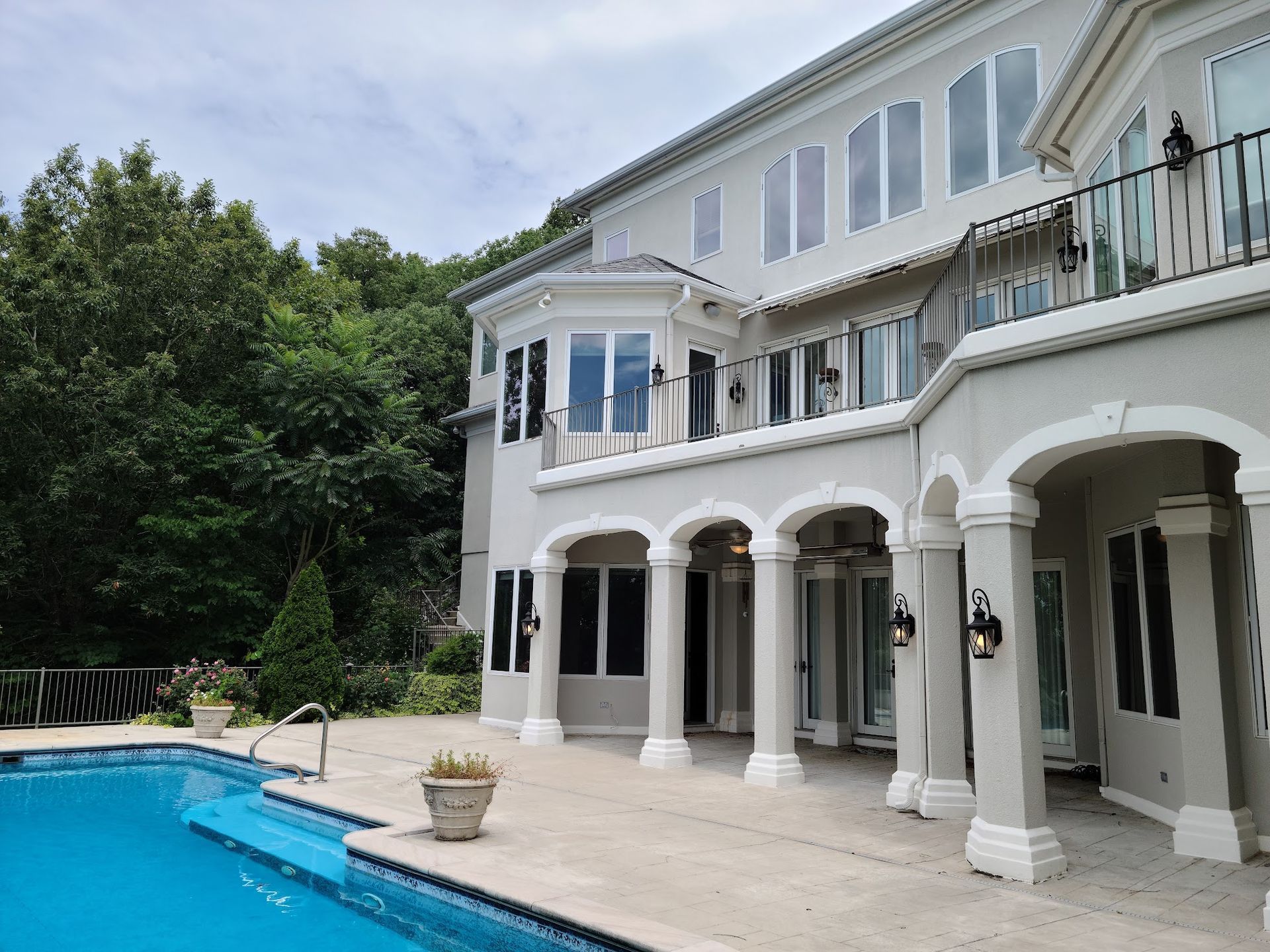 Back of a large gray mansion with a pool, balcony, and lush trees.