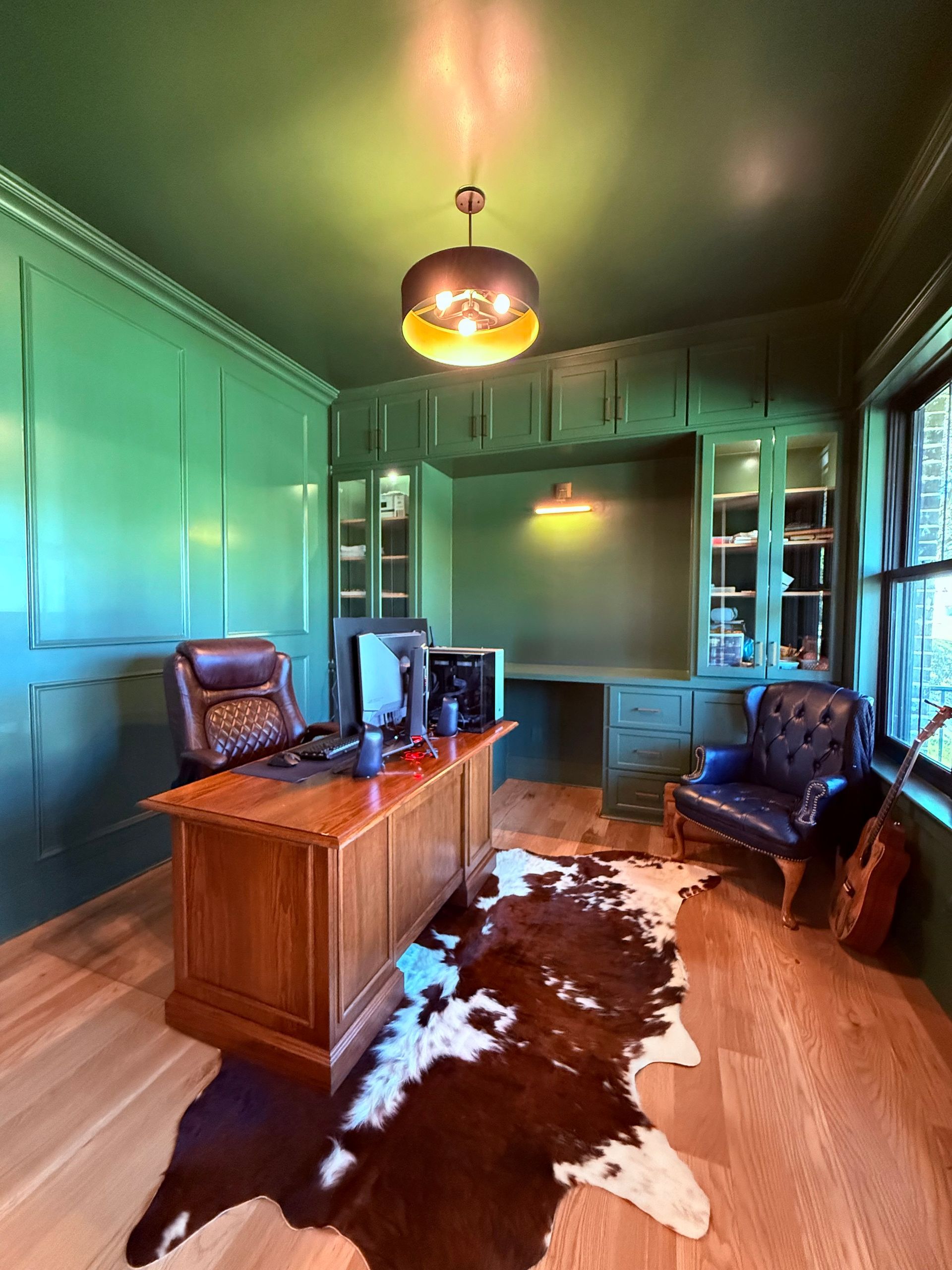 Vintage room with green walls, wooden desk, leather chairs, and cowhide rug under a hanging lamp.