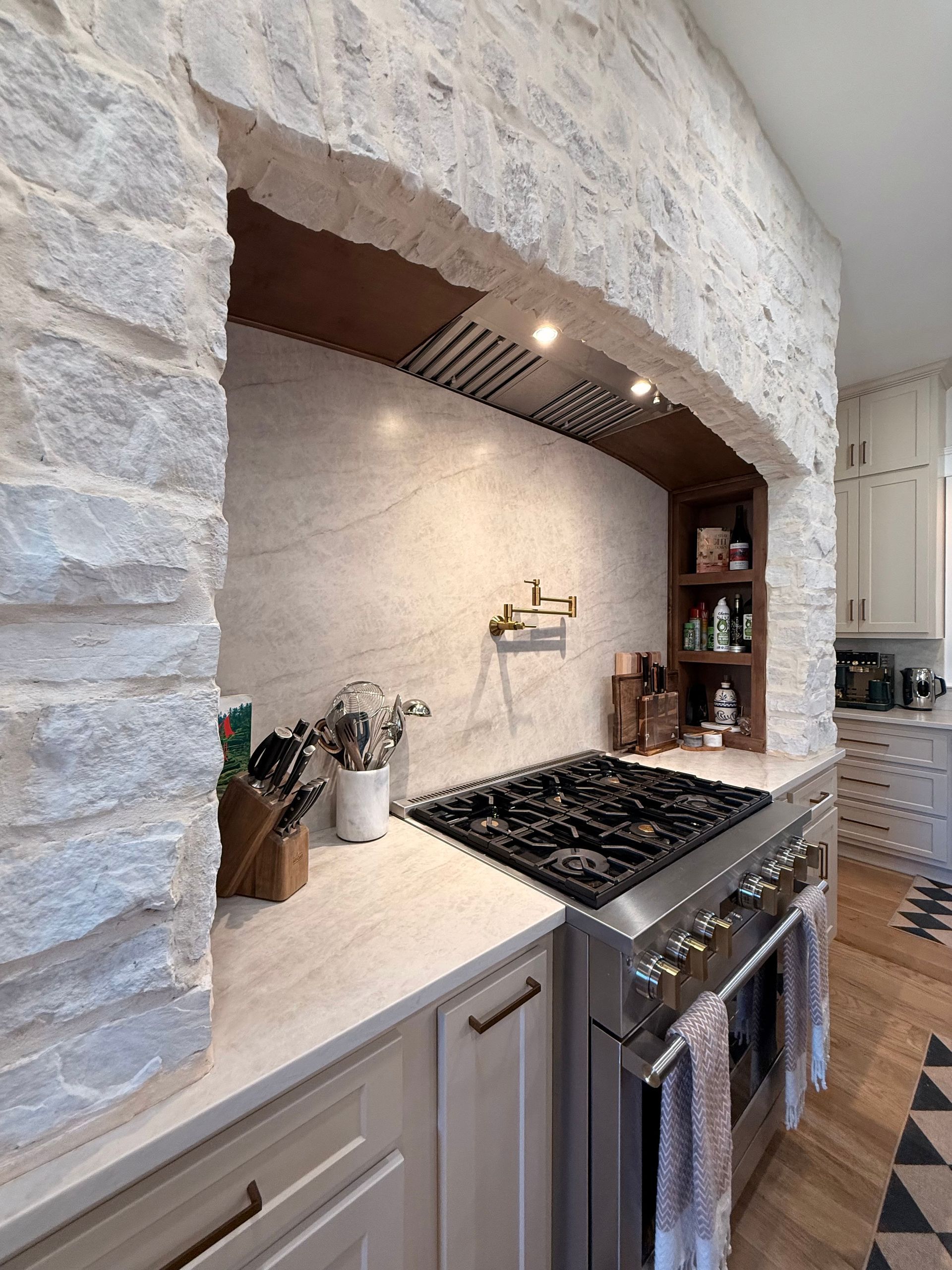 Stone-arched kitchen with white countertops, stainless stove, and warm wood shelves