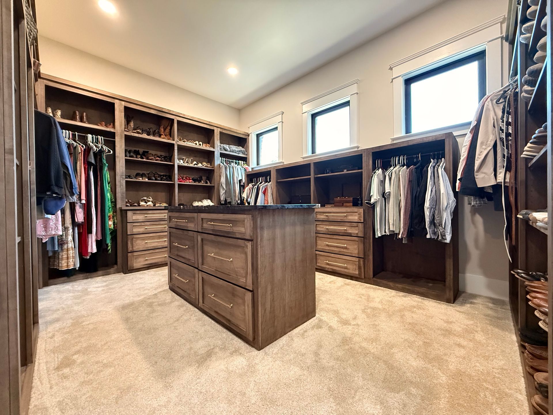 Spacious walk-in closet with wooden drawers, hanging clothes, and two windows letting in natural light