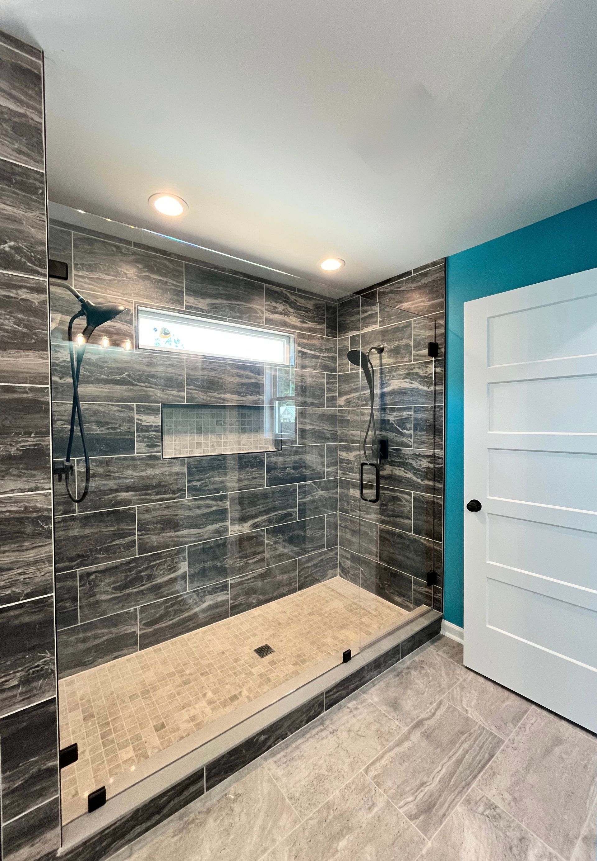 Modern bathroom with walk-in shower, gray tile walls, beige floor tile, and white door.