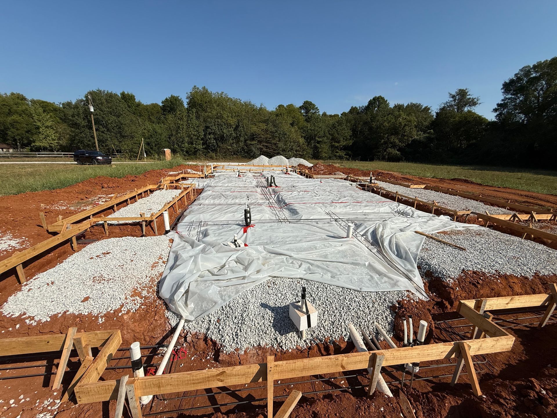 Construction site with a large lined excavation pit covered in white plastic sheeting and surrounded by dirt and lumber