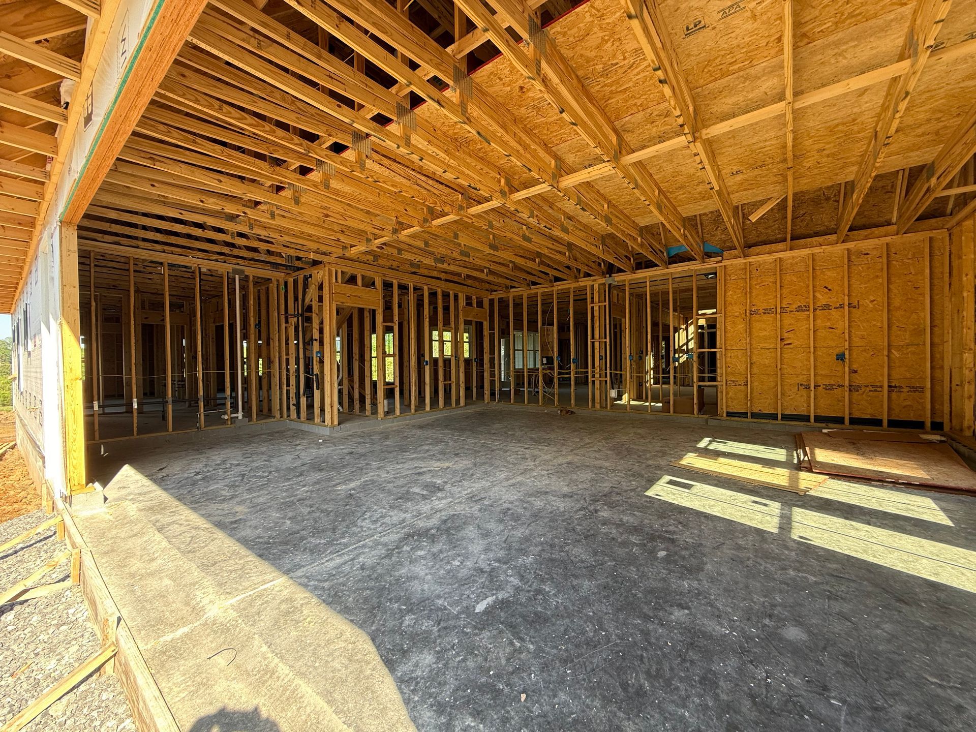 Unfinished house interior with exposed wooden framing and concrete floor, sunlight streaming through openings.