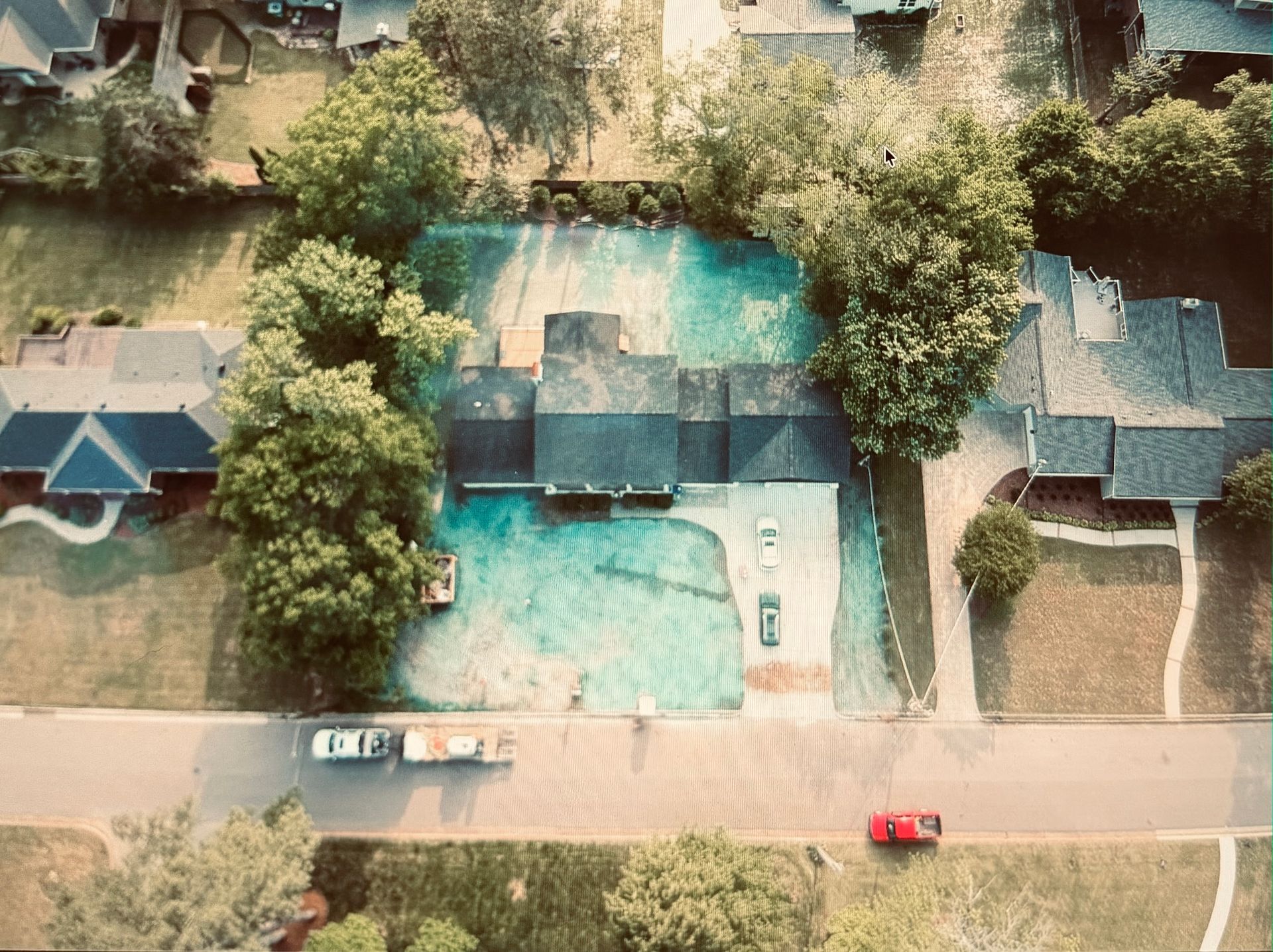 Aerial view of a house with a turquoise pool, trees, parked cars, and a red car on the street.