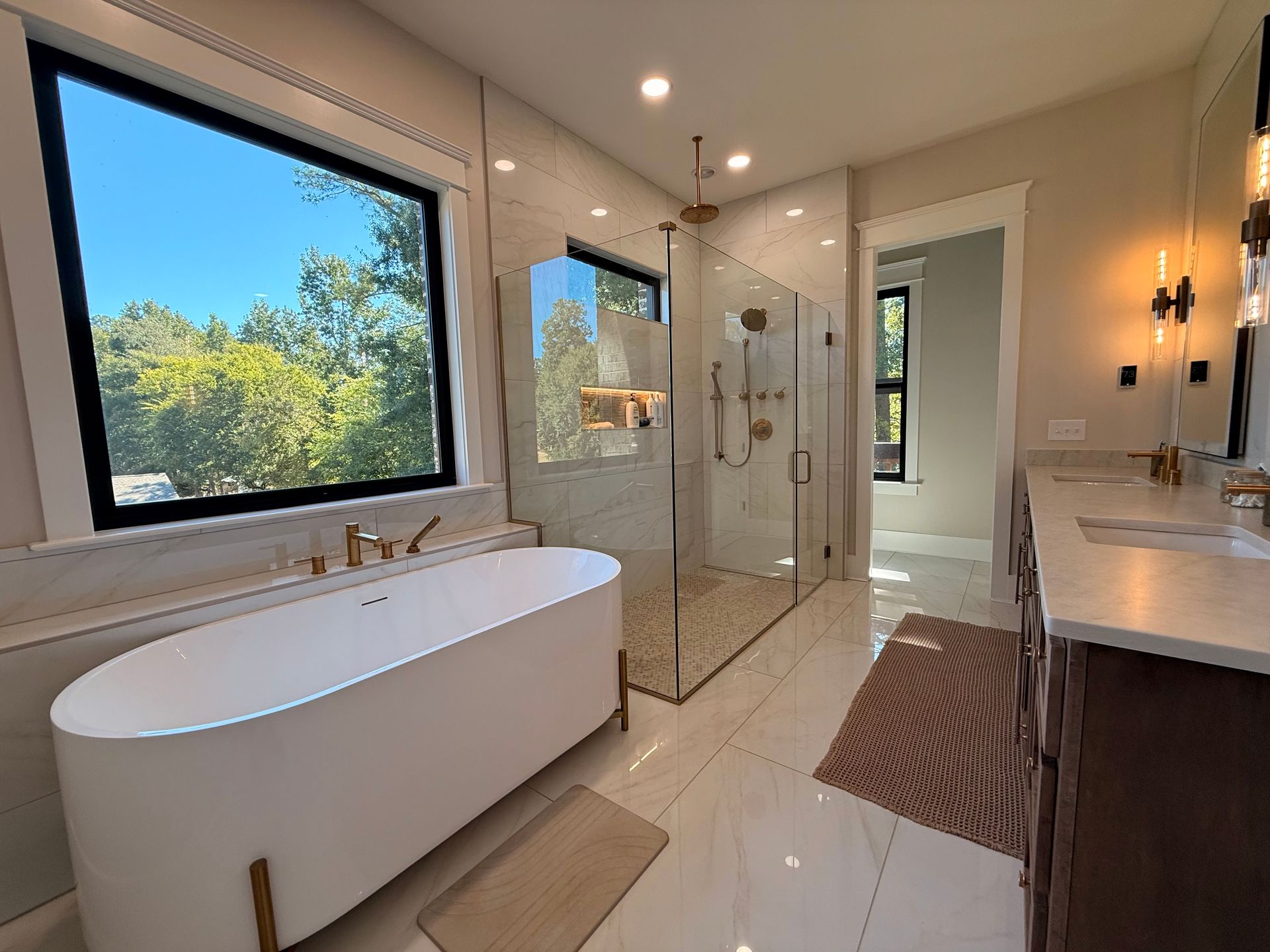 Modern bathroom with freestanding tub, glass shower, double vanity, and large window overlooking greenery