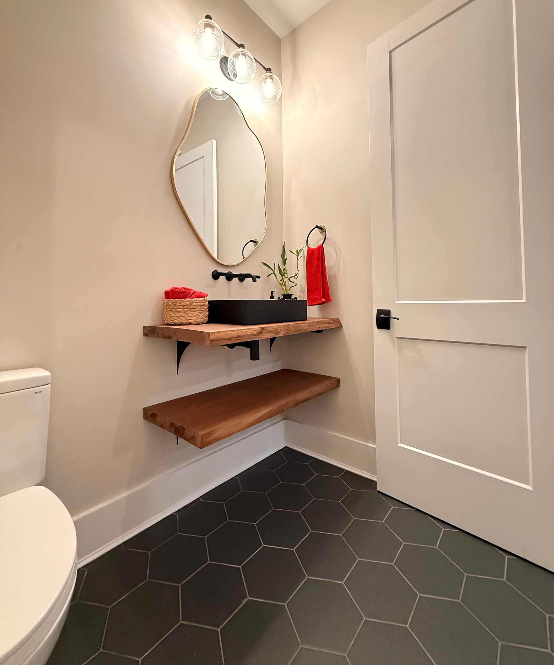 Small bathroom nook with black hex tile, wood shelves, round mirror, sink, and white door.