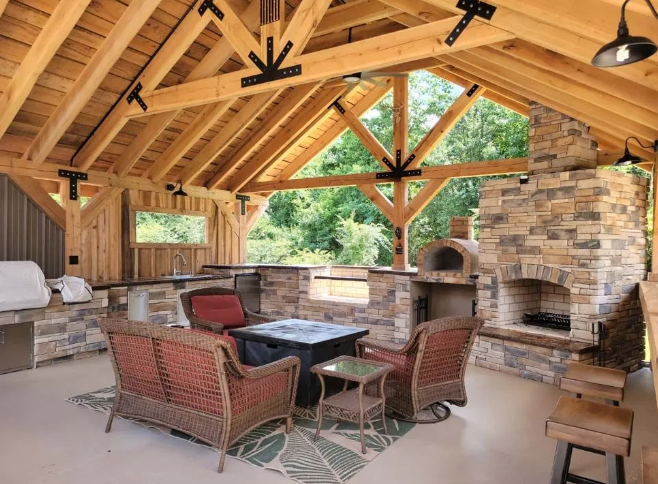 Outdoor kitchen with stone accents, fireplace, and seating under a wooden roof.